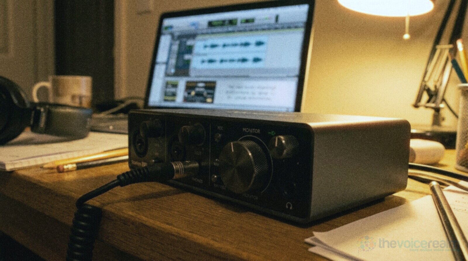 A home audio interface with a coiled headphone cable plugged into its front panel and a large monitor knob, a laptop showing a waveform behind it in lamp light © The Voice Realm