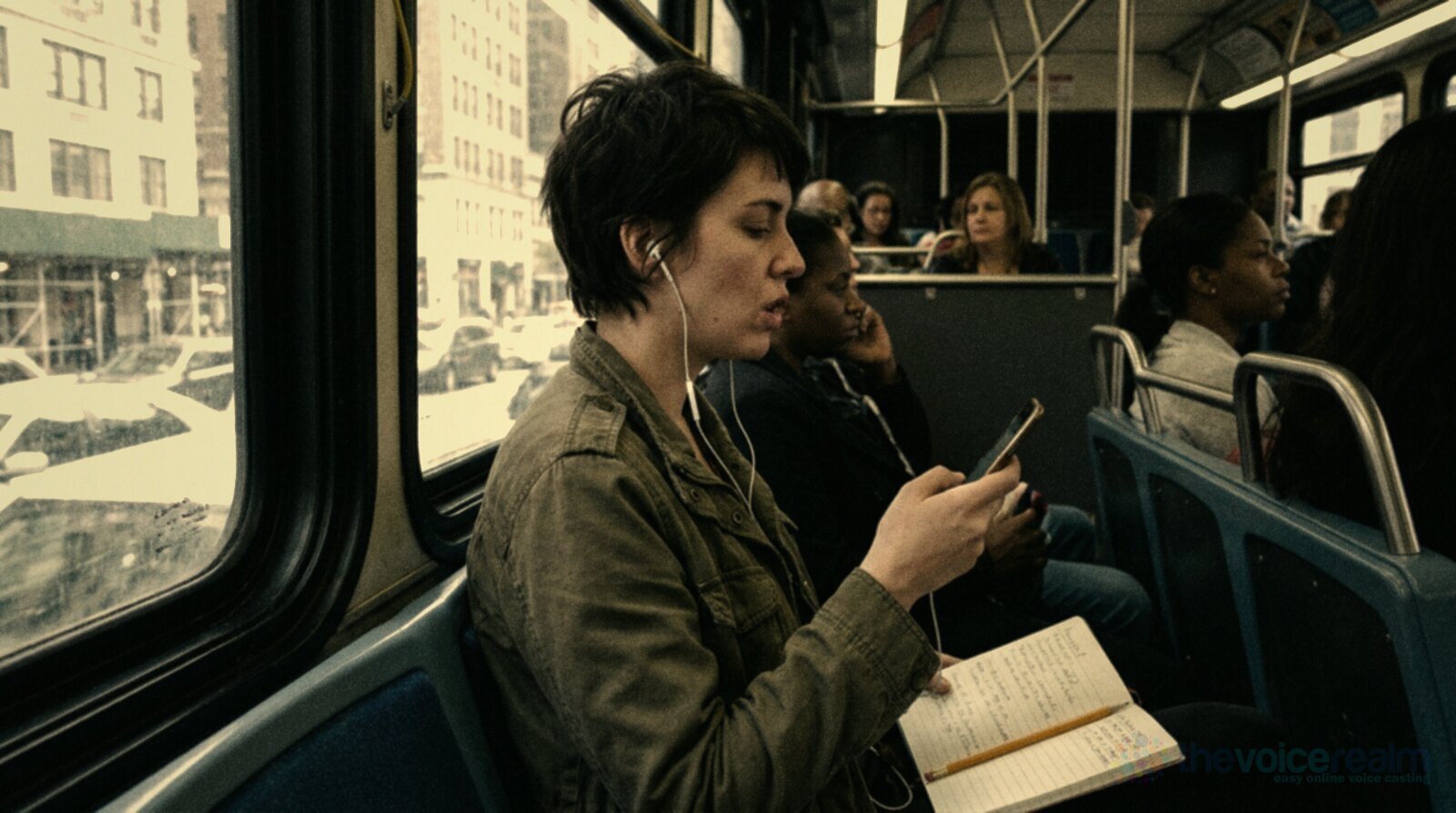 A woman with short dark hair sitting by the window on a city bus with wired earbuds in, quietly mouthing along to a recording on her phone, an open notebook and a pencil on her knee © The Voice Realm
