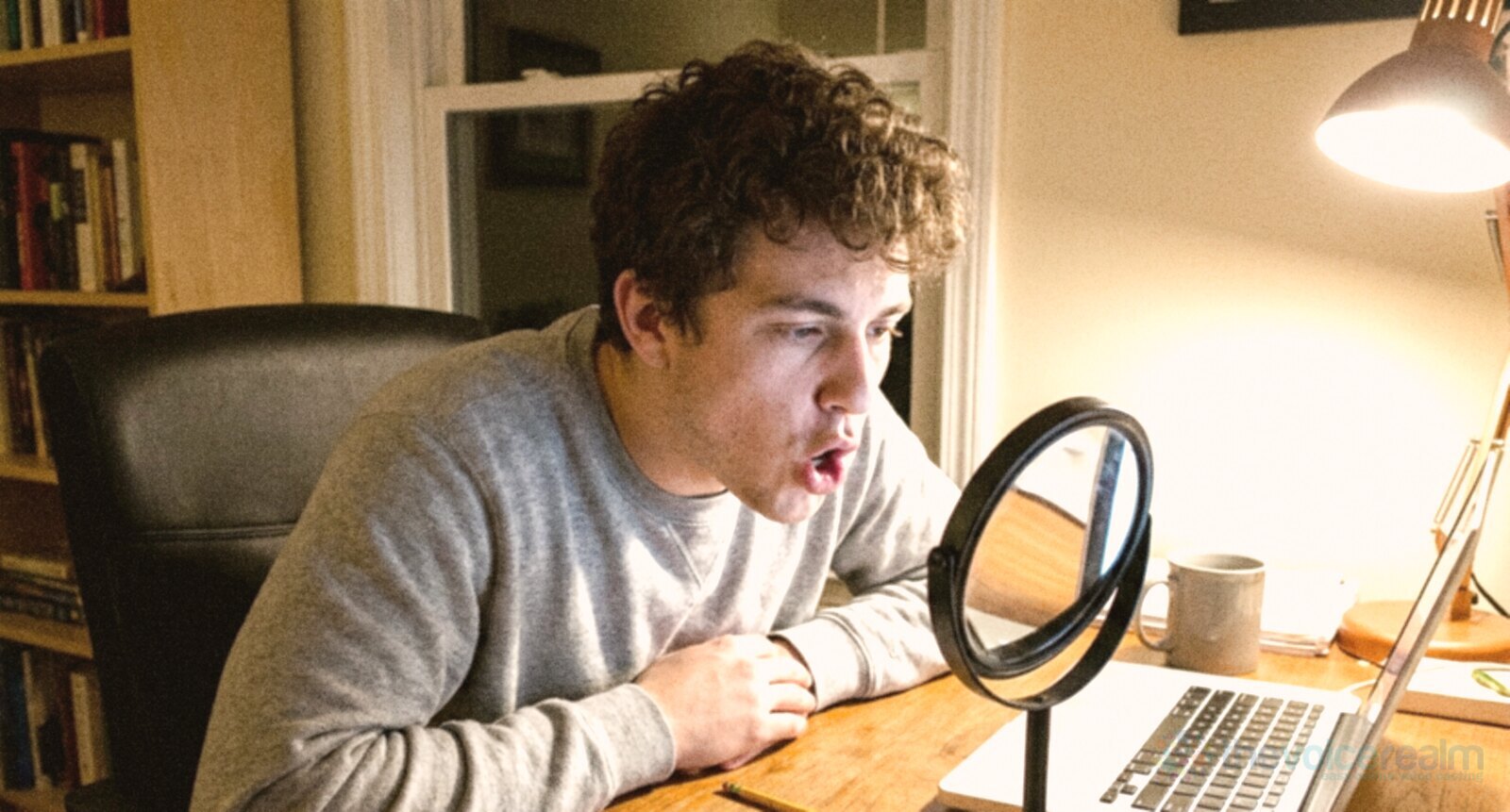 A young man with curly hair at a desk at night, shaping an open vowel with his mouth while watching himself in a small round stand mirror beside his laptop © The Voice Realm