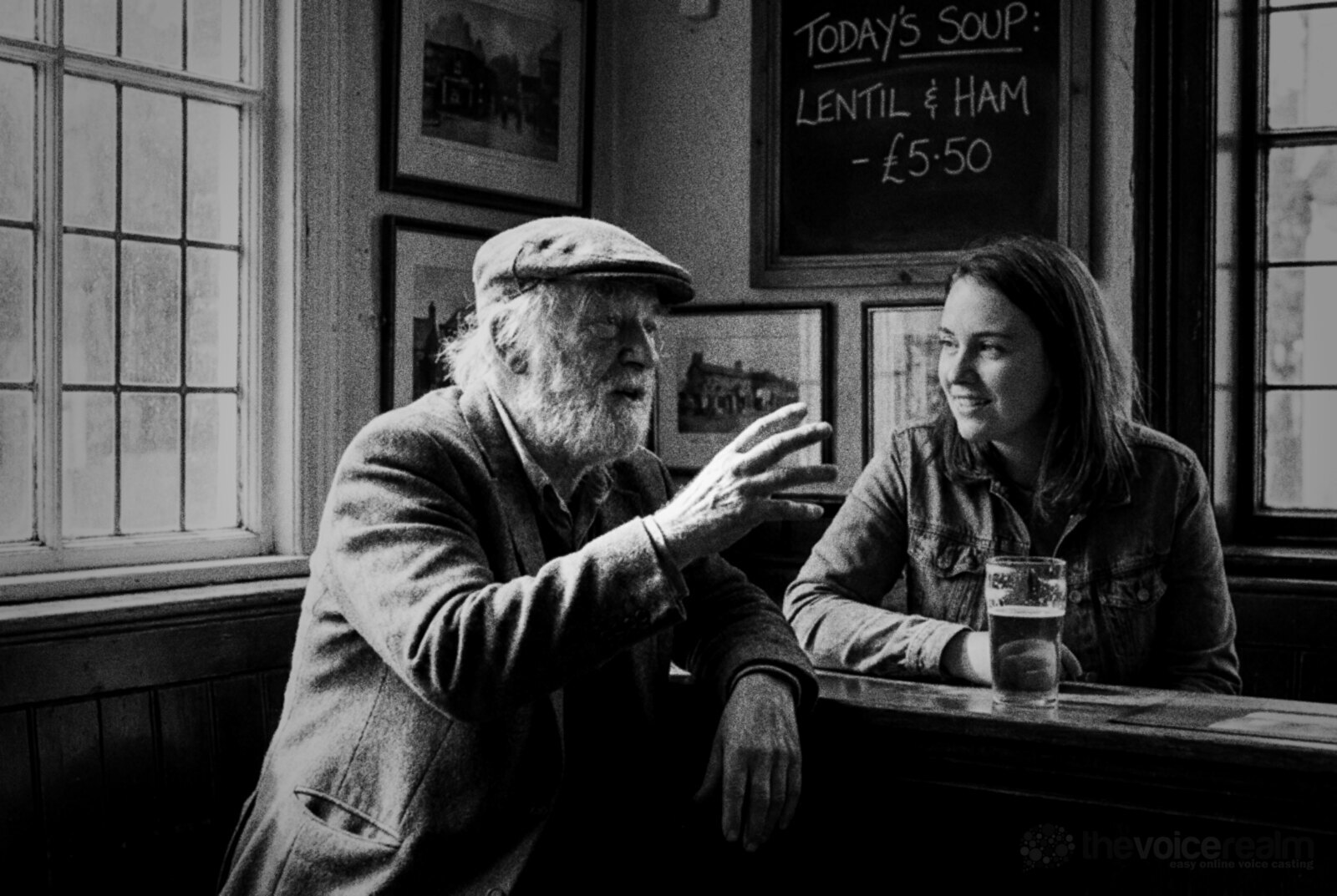 Black and white photo of an older man with a white beard and a flat cap telling a story at a pub bar while a younger woman in a denim jacket listens closely © The Voice Realm