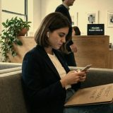 A young woman in a navy jacket with earbuds in, waiting on a grey sofa in a bright reception area with a folder on her knees, checking her phone, a plant and framed prints behind her © The Voice Realm