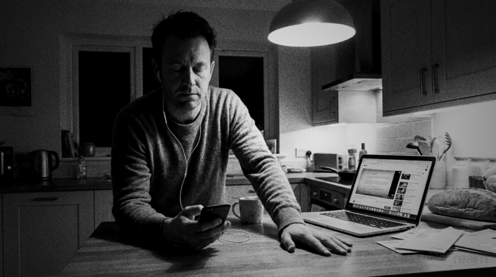 Black and white photo of a man in a jumper standing at his kitchen counter at night listening on wired earbuds to his phone with his eyes lowered, a laptop open beside him © The Voice Realm