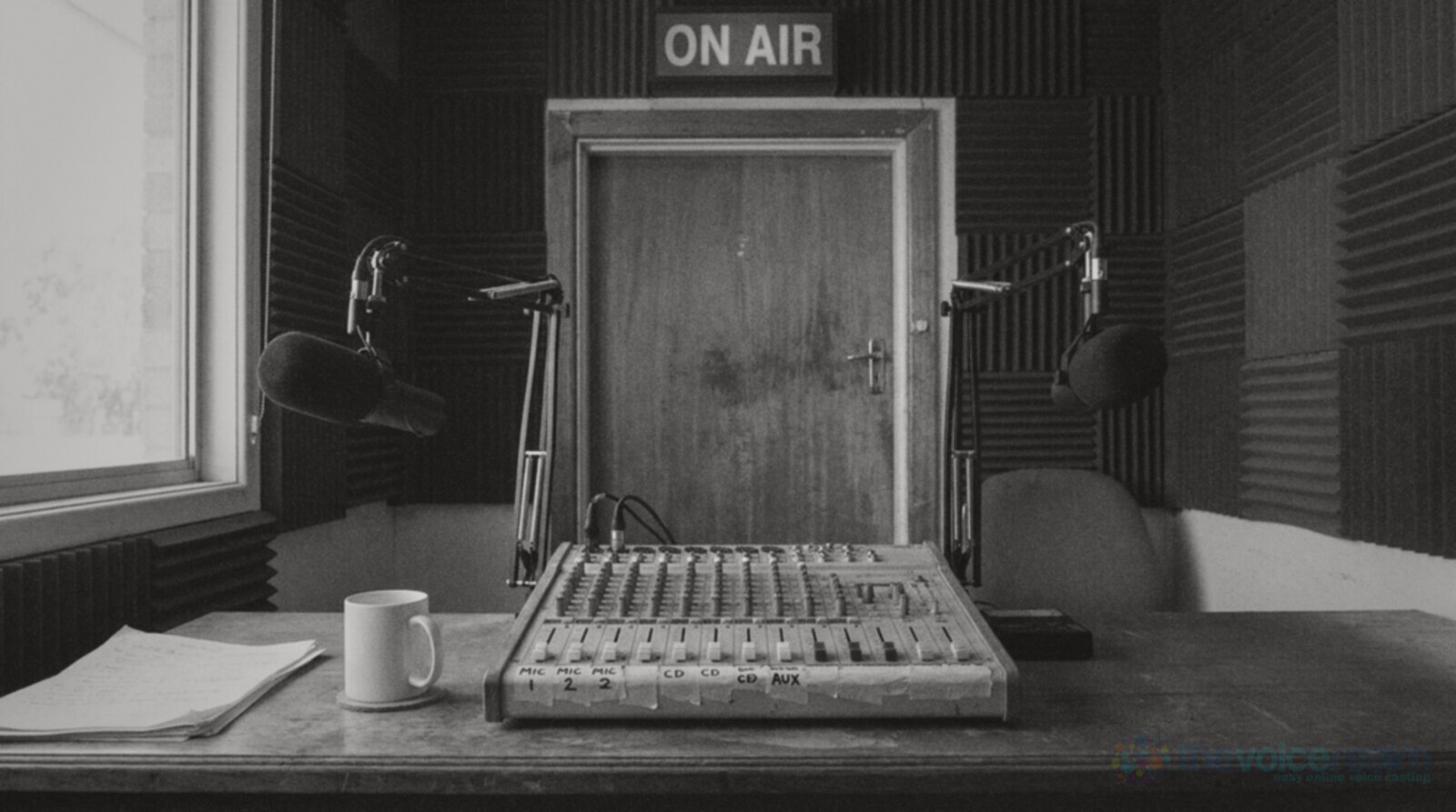 A small community radio studio with two microphones on boom arms, a mixing desk labelled with masking tape, a red On Air light above the door and a plain white mug on the desk © The Voice Realm