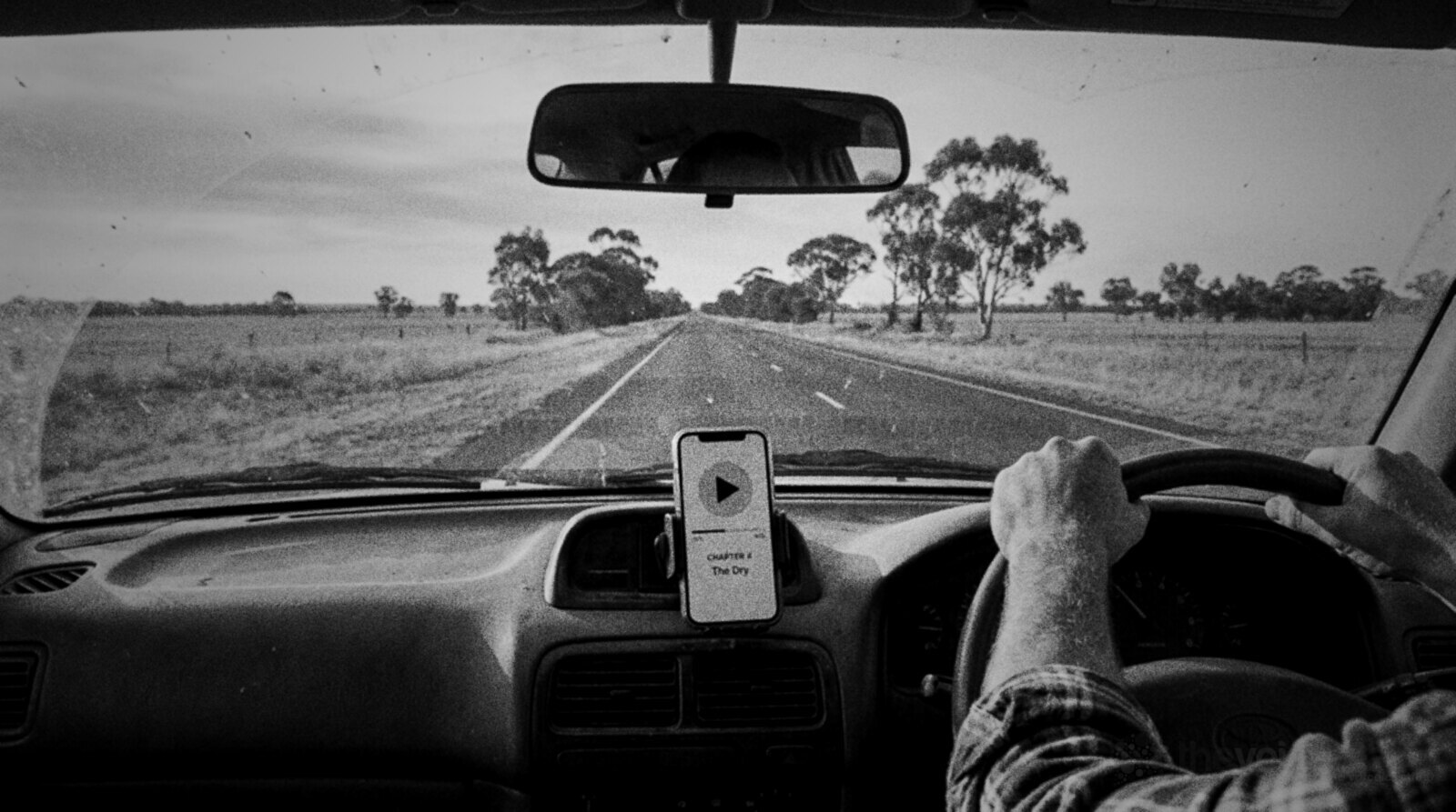 Black and white view from inside a car on a long straight country highway through flat paddocks and gum trees, the driver's hands on the wheel and a phone in a dashboard cradle playing an audiobook © The Voice Realm