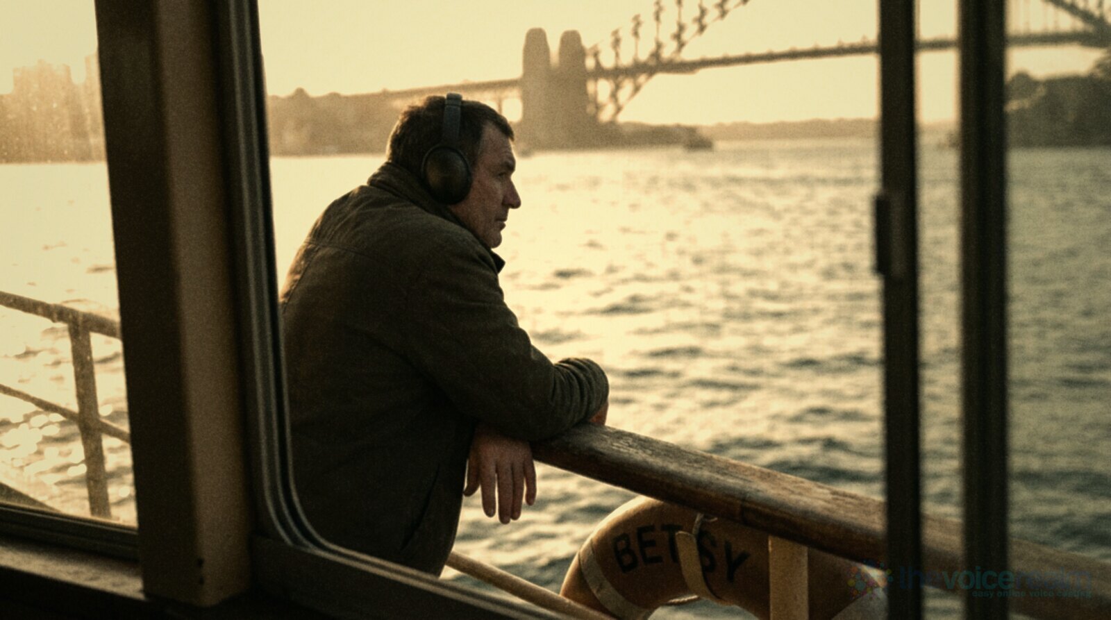 A man in a dark jacket and over-ear headphones leans on the timber rail of a Sydney ferry at golden hour, looking out across the water with the Harbour Bridge soft in the background © The Voice Realm