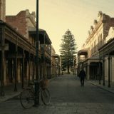 A quiet heritage street in the West End of Fremantle early in the morning, limestone and red brick shopfronts with iron lace verandahs, a Norfolk Island pine at the end of the street, a bicycle leaning on a post and one person walking away, in warm faded film colours © The Voice Realm