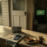 Morning in the kitchen of an old Queensland weatherboard house: a small portable radio, a phone on a tea towel, toast and a mug of tea on the laminate bench, louvre windows and a security screen door, and through the doorway a television in the lounge room showing a cricket match, in warm faded film colours © The Voice Realm