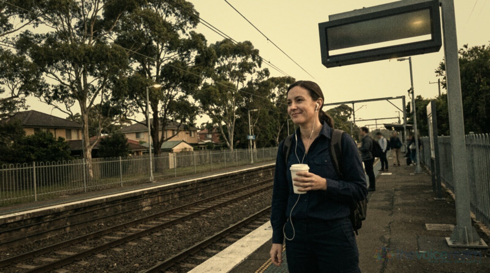 A commuter in a navy work shirt waiting on a Sydney suburban railway platform in early morning light, white wired earbuds in, holding a takeaway coffee, gum trees and red brick houses beyond the fence © The Voice Realm