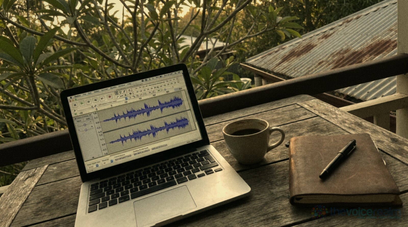 An open laptop showing an audio waveform on a weathered timber table on the back verandah of a Queenslander, a cup of coffee and a leather notebook beside it, frangipani and a rusty corrugated iron roof behind © The Voice Realm