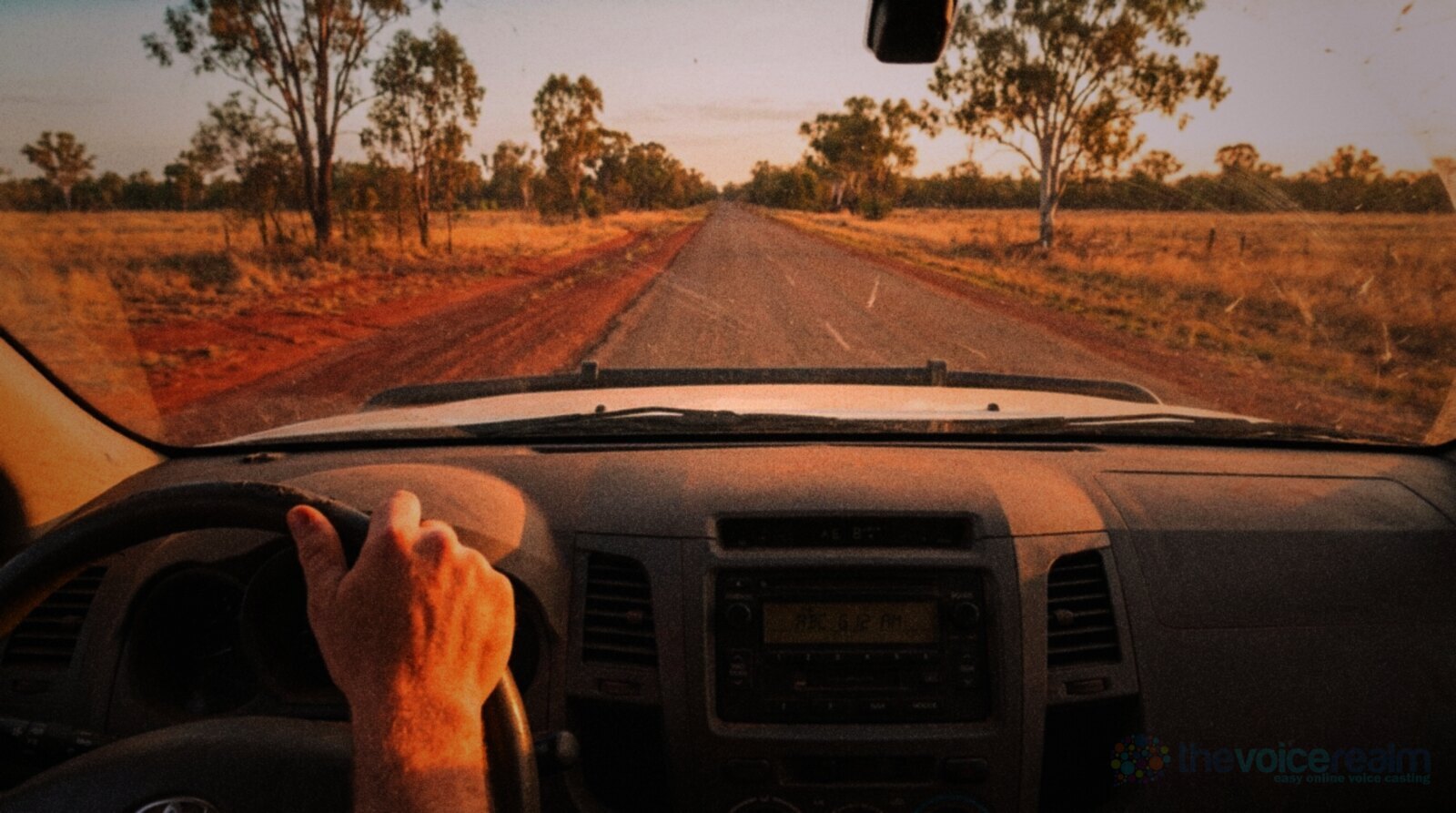 View from the driver's seat of a dusty ute on a straight country road in rural Queensland at golden hour, red dirt verges and gum trees ahead, the car radio glowing on the dashboard © The Voice Realm