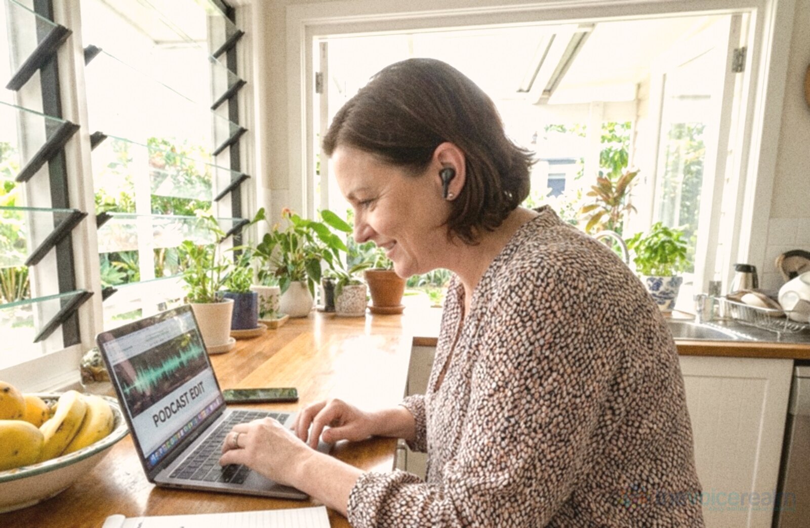 A small business owner at the kitchen bench of a Brisbane house listening through earbuds to audio on her laptop, louvre windows, pot plants and a bowl of mangoes beside her © The Voice Realm