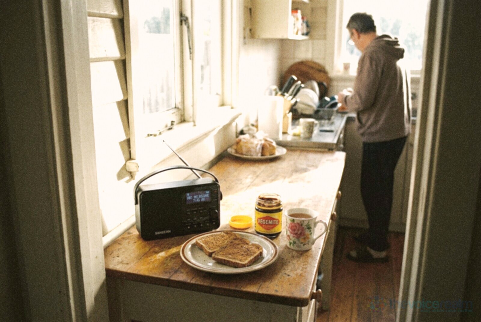 A weatherboard kitchen in the morning with a small digital radio on the timber bench beside toast, a jar of Vegemite and a floral mug of tea, a person washing up in the background © The Voice Realm
