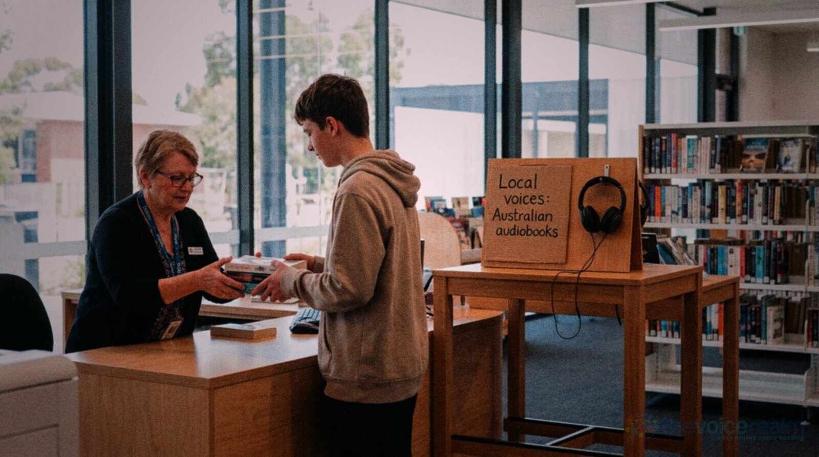 A librarian handing books to a teenager at the returns desk of a bright Australian public library, beside a display stand with headphones and a handwritten sign reading Local voices: Australian audiobooks © The Voice Realm