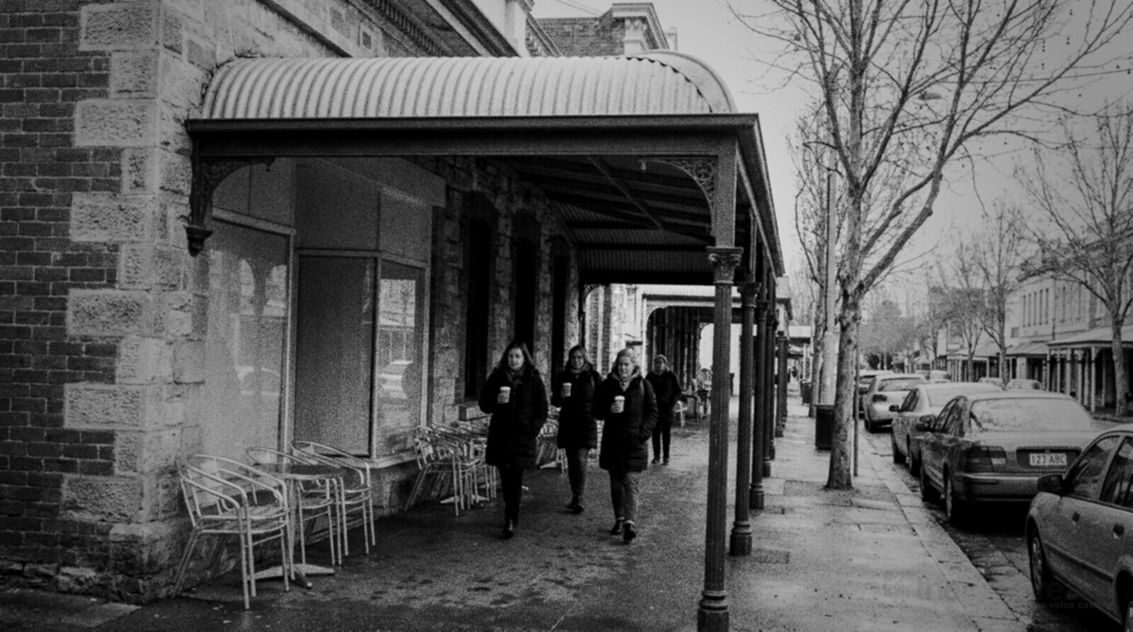 Black and white photo of a heritage street in the East End of Adelaide on an overcast morning, sandstone and red brick shopfronts under a curved iron verandah on green posts, empty cafe chairs on the footpath and three women walking with takeaway coffee past bare plane trees © The Voice Realm