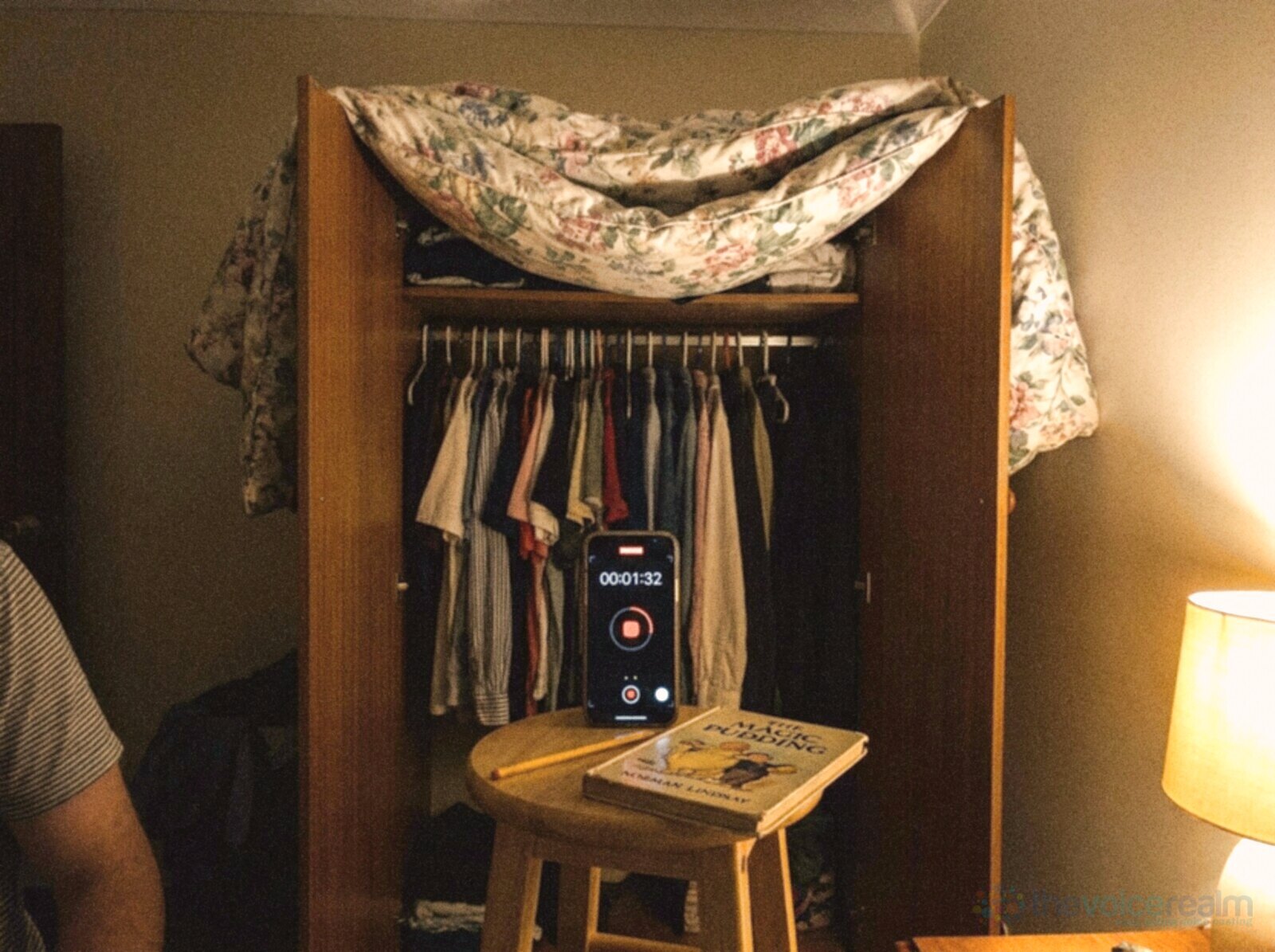 An open wardrobe in a bedroom at night turned into a recording booth, a floral doona thrown over the doors, a smartphone recording on a stool in front of the hanging clothes beside a copy of The Magic Pudding and a pencil, a bedside lamp glowing, in bright phone snapshot colours © The Voice Realm