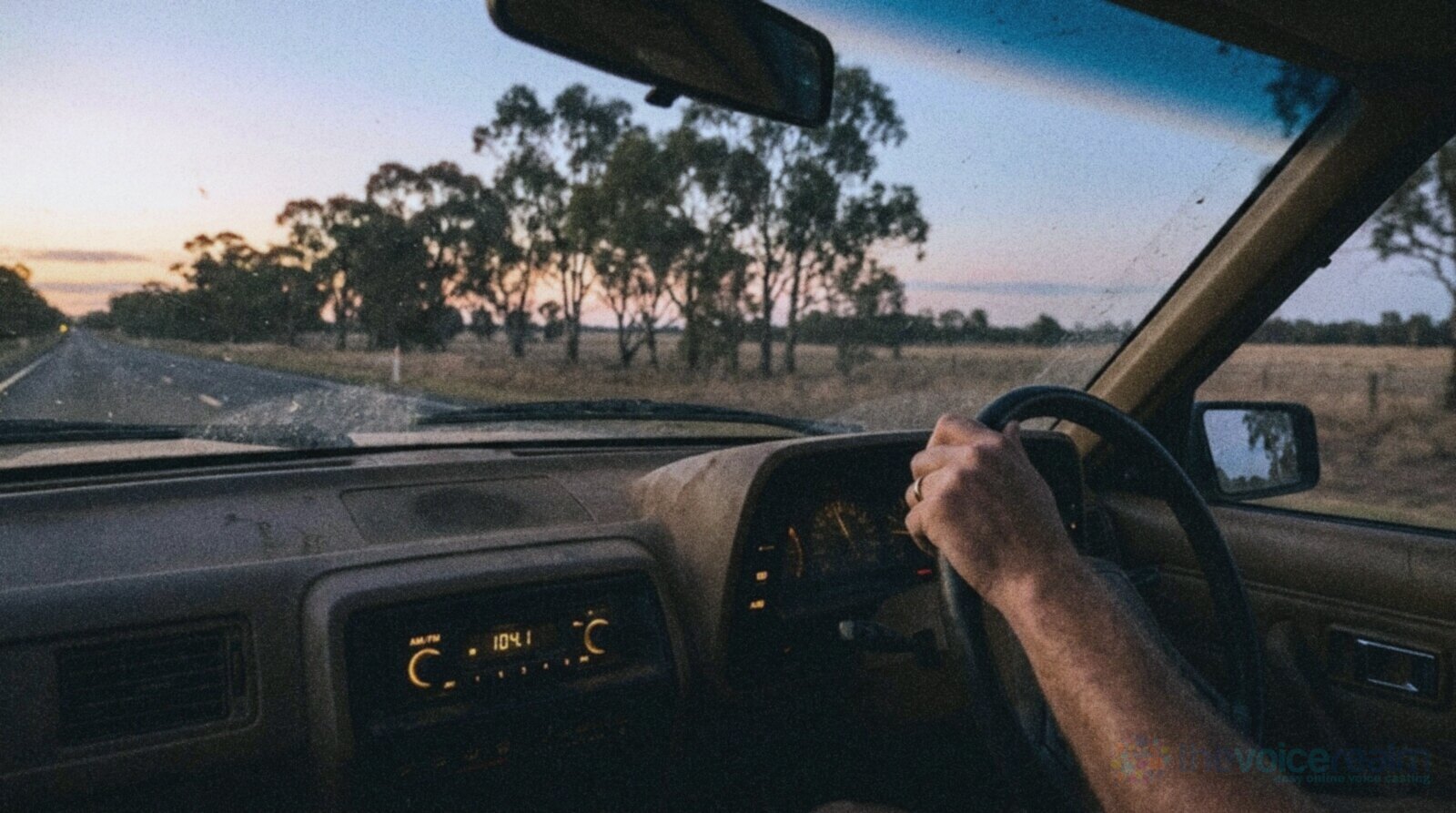 The view from the passenger seat of a dusty old car on a straight country highway in New South Wales at dusk, the dashboard radio glowing amber, the driver's hand on the wheel, paddocks and gum trees under a pink and orange sky, in cool grainy film colours © The Voice Realm