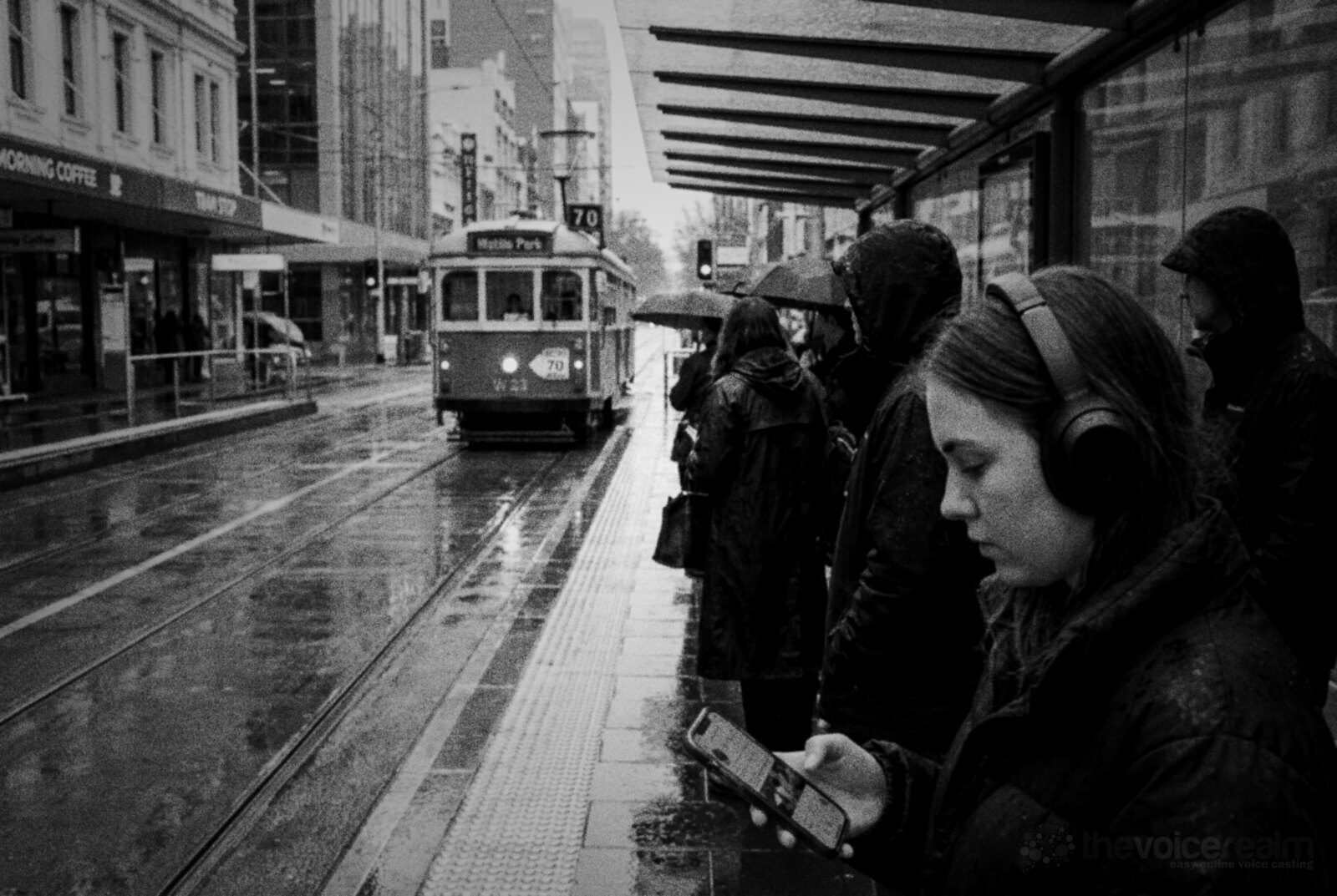 Black and white photo of commuters waiting at a Melbourne tram stop in the rain, a young woman in over-ear headphones looking at her phone as a green and cream tram approaches © The Voice Realm