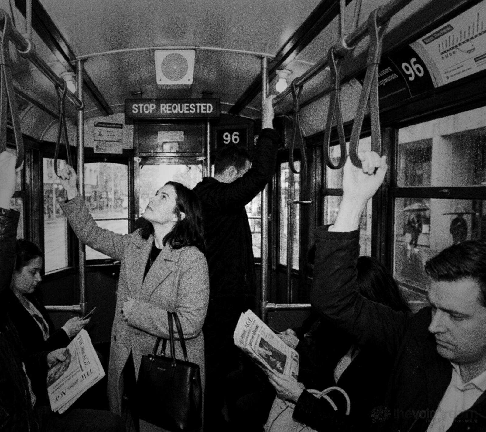 Black and white photo of commuters inside a Melbourne tram on a rainy morning, a woman holding a strap looks up at the ceiling speaker under a Stop Requested sign © The Voice Realm