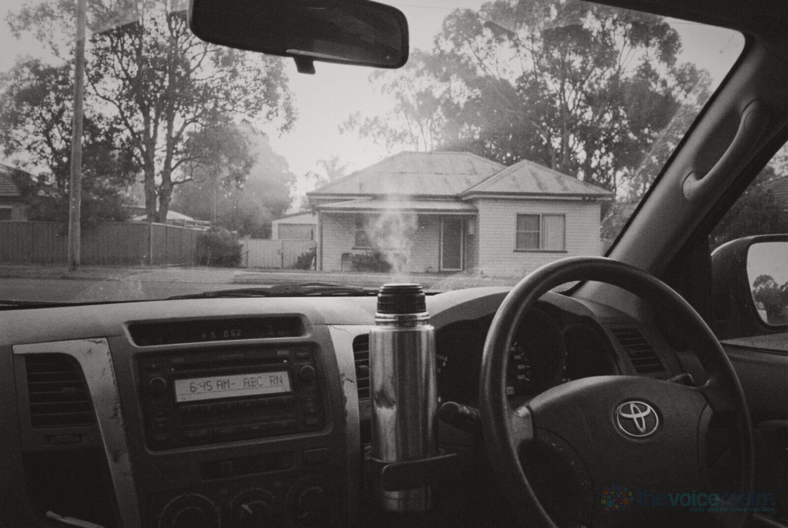 View from the driver's seat of a ute parked on a quiet Australian suburban street at sunrise, a steaming thermos in the cup holder and the radio display glowing © The Voice Realm