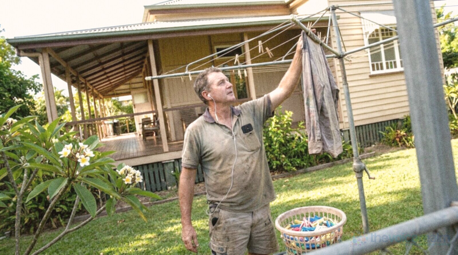 A man hanging washing on a rotary clothes line in the backyard of a Queenslander with a timber verandah, one earbud in and his phone in his shirt pocket, a frangipani tree in flower beside him © The Voice Realm
