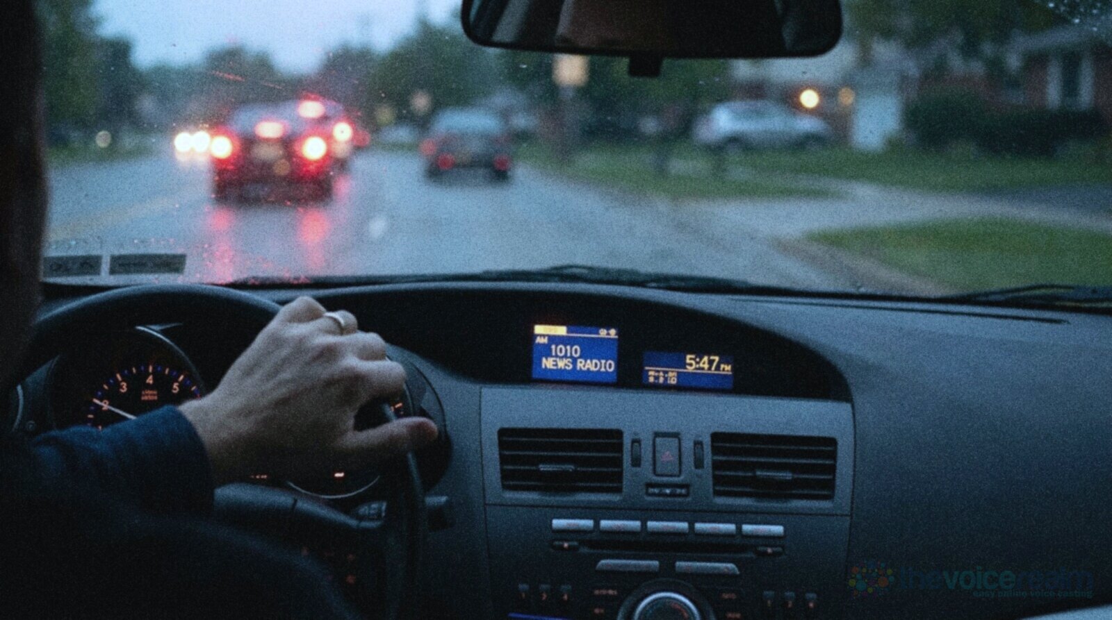 View from inside a car at dusk in the rain, the driver's hand on the wheel, brake lights of traffic ahead and the dashboard radio display lit up reading AM 1010 NEWS RADIO and 5:47 PM © The Voice Realm