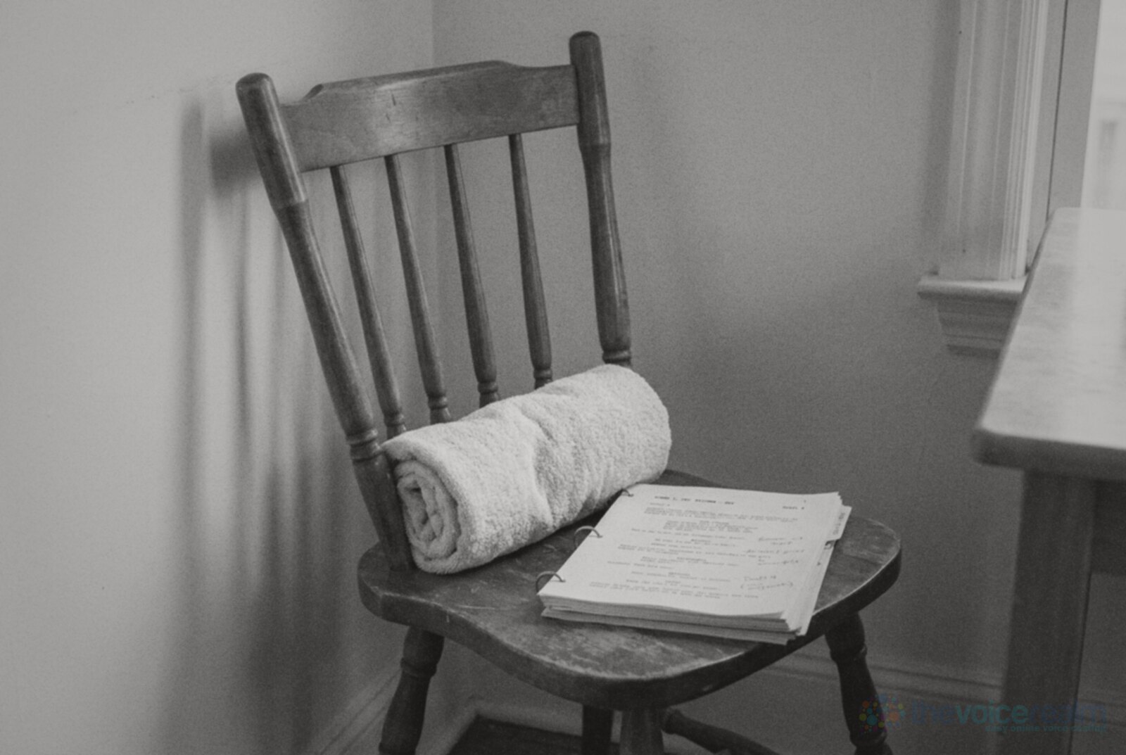 A plain wooden spindle-back chair in the corner of a white room with a rolled towel tucked against the lower back rest and a ring-bound script on the seat, black and white photograph © The Voice Realm