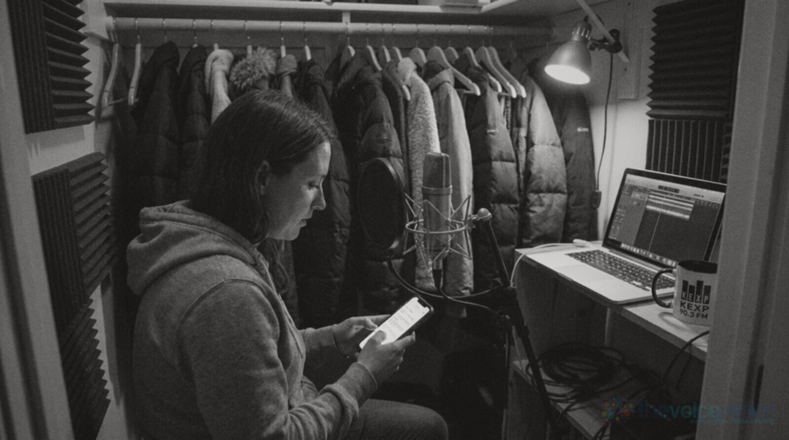 Black and white photo of a woman in a hoodie recording at night in a walk-in closet lined with coats and foam panels, reading a short script from her phone in front of a condenser microphone with a pop filter, a laptop open on a shelf beside her © The Voice Realm