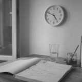 Black and white photo of a kitchen table by an open window, a round wall clock above, a printed script, a glass of water, a jar of honey and a steel thermos flask © The Voice Realm