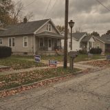 A quiet suburban street in late October with fallen leaves on the lawns, campaign signs reading VOTE and EARLY VOTING NOV 3 pushed into the grass outside clapboard houses, a mailbox and a pumpkin on a porch under a grey sky © The Voice Realm