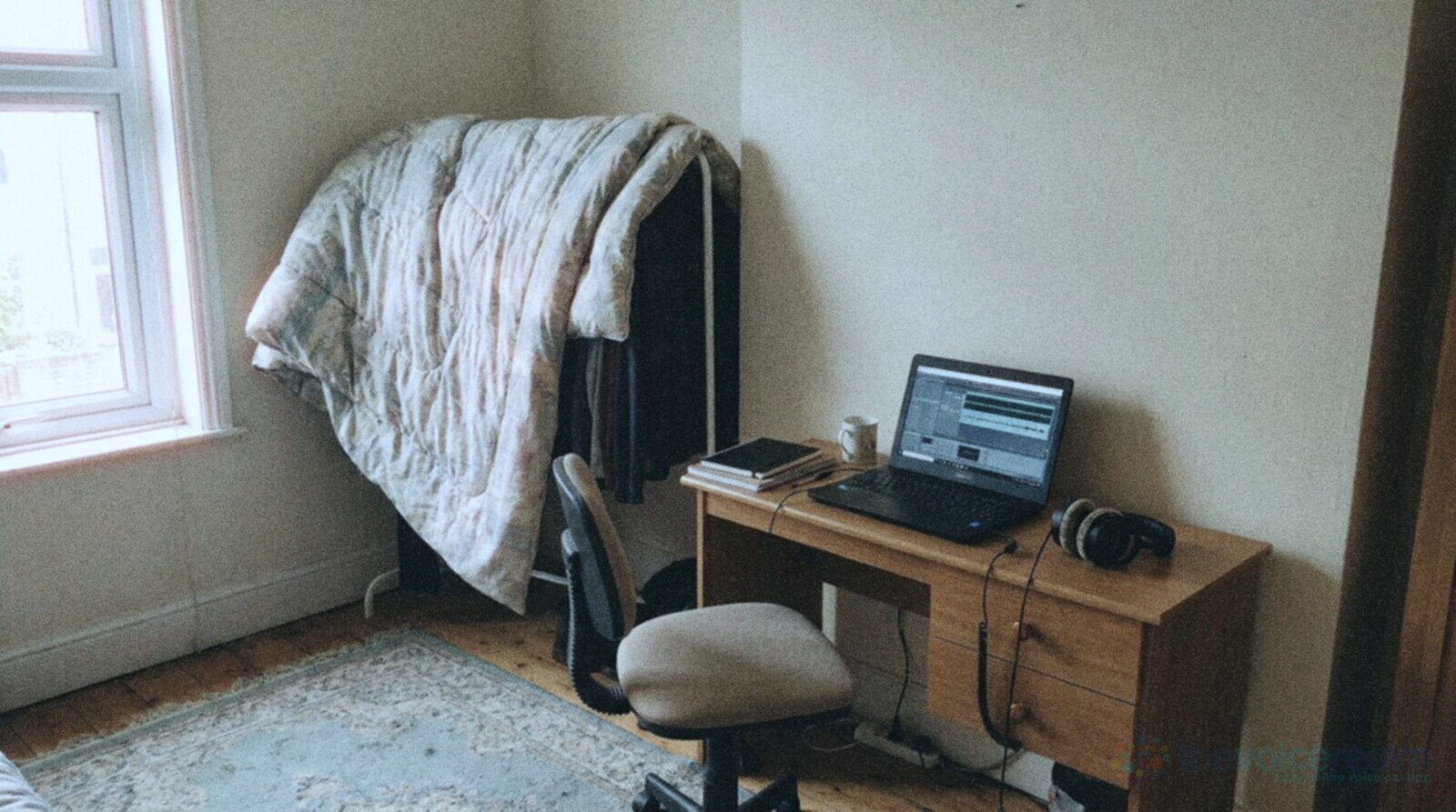 A small spare bedroom turned into a home recording corner, with a plain desk, a laptop and headphones, a duvet hung over a clothes rack behind the chair, a rug on the floor and daylight from a side window © The Voice Realm