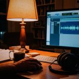 A home office desk at night lit by a table lamp, a hand on the mouse in front of a monitor showing a single blue spoken word waveform in audio editing software, with closed-back headphones, a keyboard, script pages and a mug of tea, in rich warm film colours © The Voice Realm