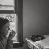 A woman in a knitted jumper sneezes into a tissue beside an open sash window on a bright autumn morning, trees with turning leaves outside, closed-back headphones and stacks of script pages on the desk beside her, black and white photograph © The Voice Realm