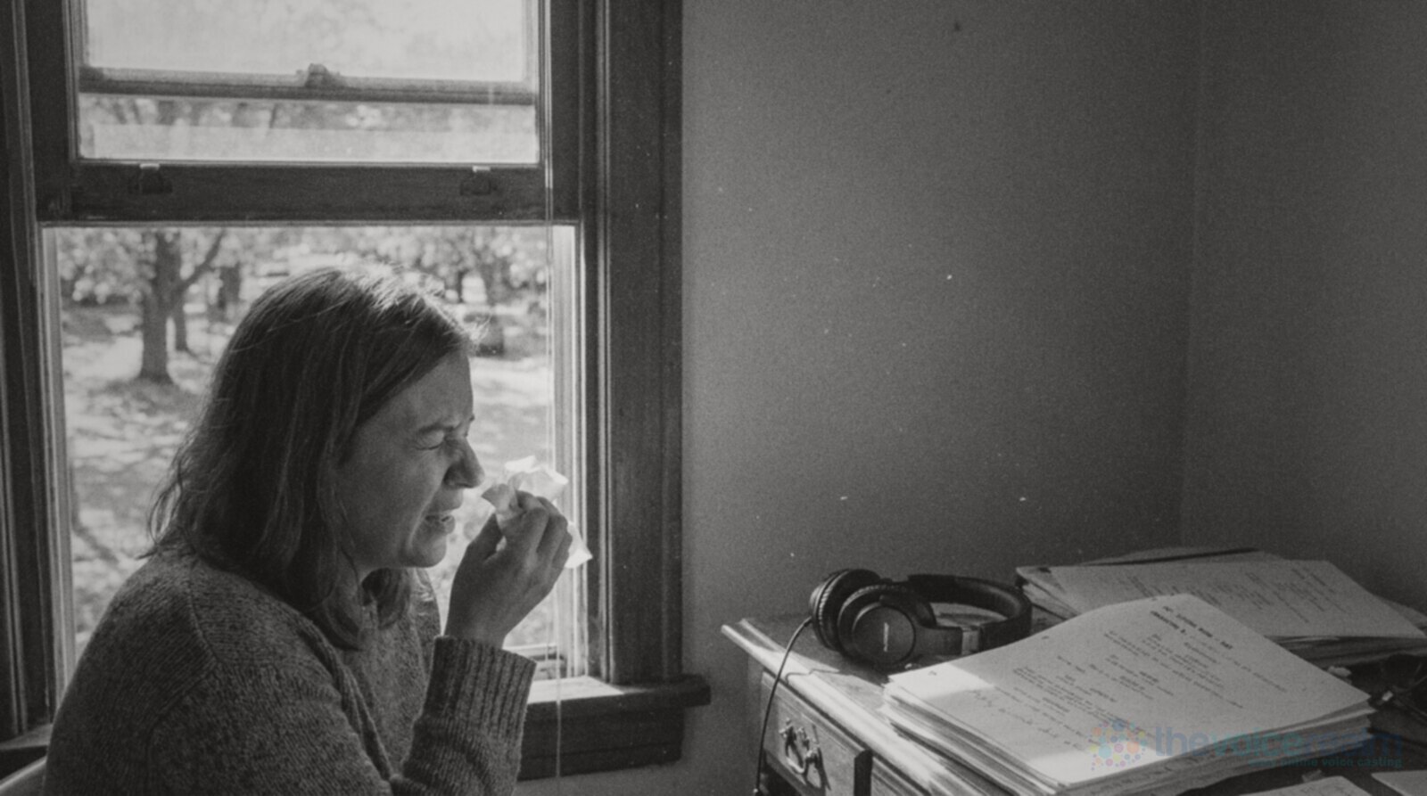A woman in a knitted jumper sneezes into a tissue beside an open sash window on a bright autumn morning, trees with turning leaves outside, closed-back headphones and stacks of script pages on the desk beside her, black and white photograph © The Voice Realm