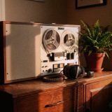 A vintage reel-to-reel tape recorder with two clear tape reels on a wooden sideboard in a sunny living room, closed-back headphones with a coiled cable beside it, a potted fern and shelves of books, in punchy warm film colours © The Voice Realm