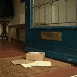 The inside of a blue front door with a brass letter slot, envelopes and a small parcel lying on a coir doormat on a red tiled hallway floor, in soft faded warm film colours © The Voice Realm