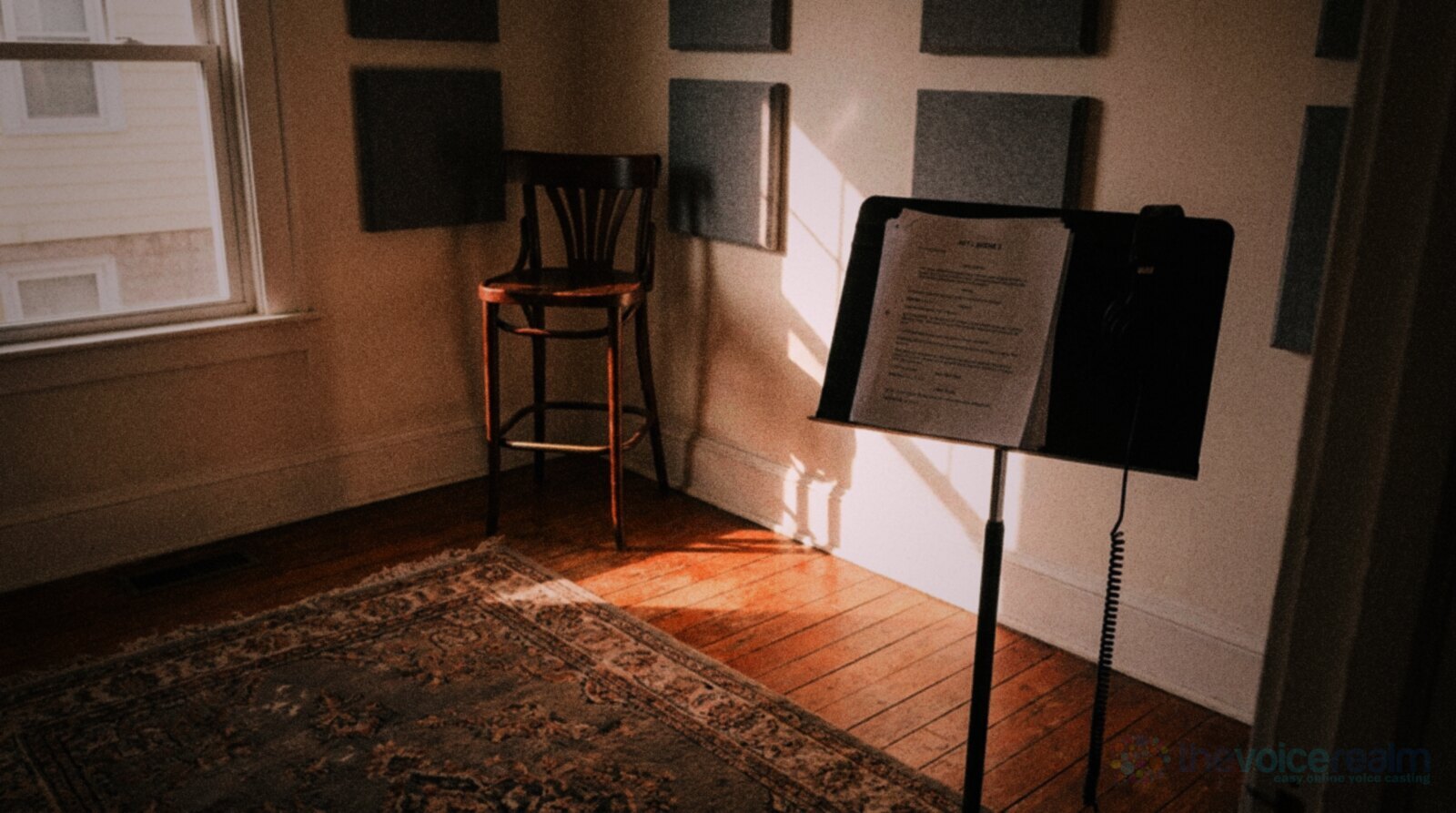 An empty spare room set up for recording in late afternoon sun: grey acoustic panels on the walls, a tall bentwood bar stool in the corner, a black music stand holding script pages with headphones hanging from it and a patterned rug on wooden floorboards, in punchy warm film colours © The Voice Realm