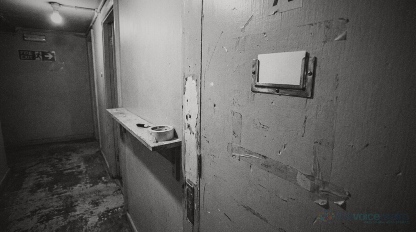 Black and white photo of a scuffed backstage dressing room door with an empty brass nameplate holding a blank card, and a roll of masking tape and a marker on a shelf beside it © The Voice Realm