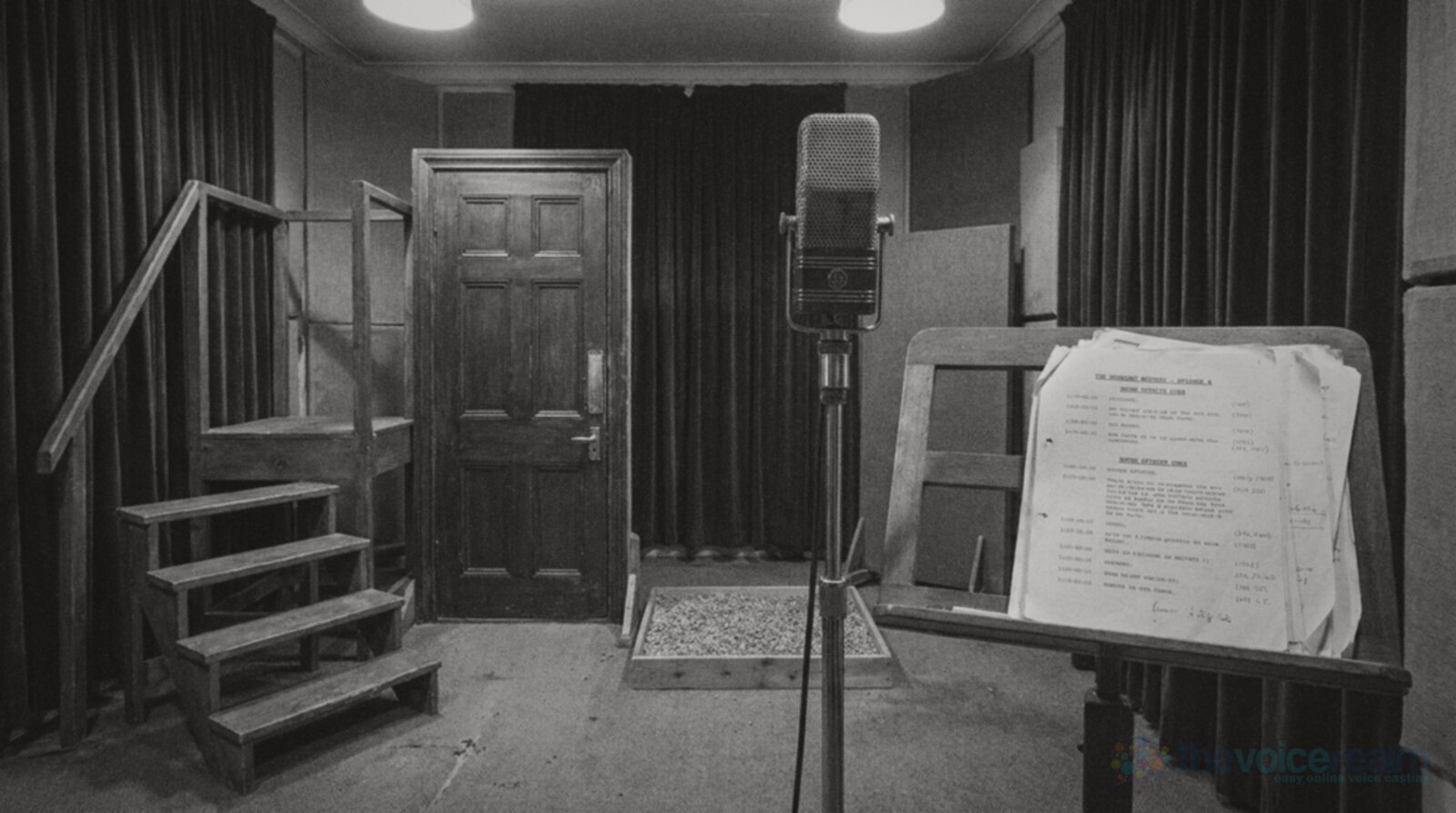 Black and white photo of an empty radio drama studio: a wooden practice staircase, a free-standing panelled door, a tray of gravel on the floor, a vintage microphone on a stand and a music stand of script pages © The Voice Realm