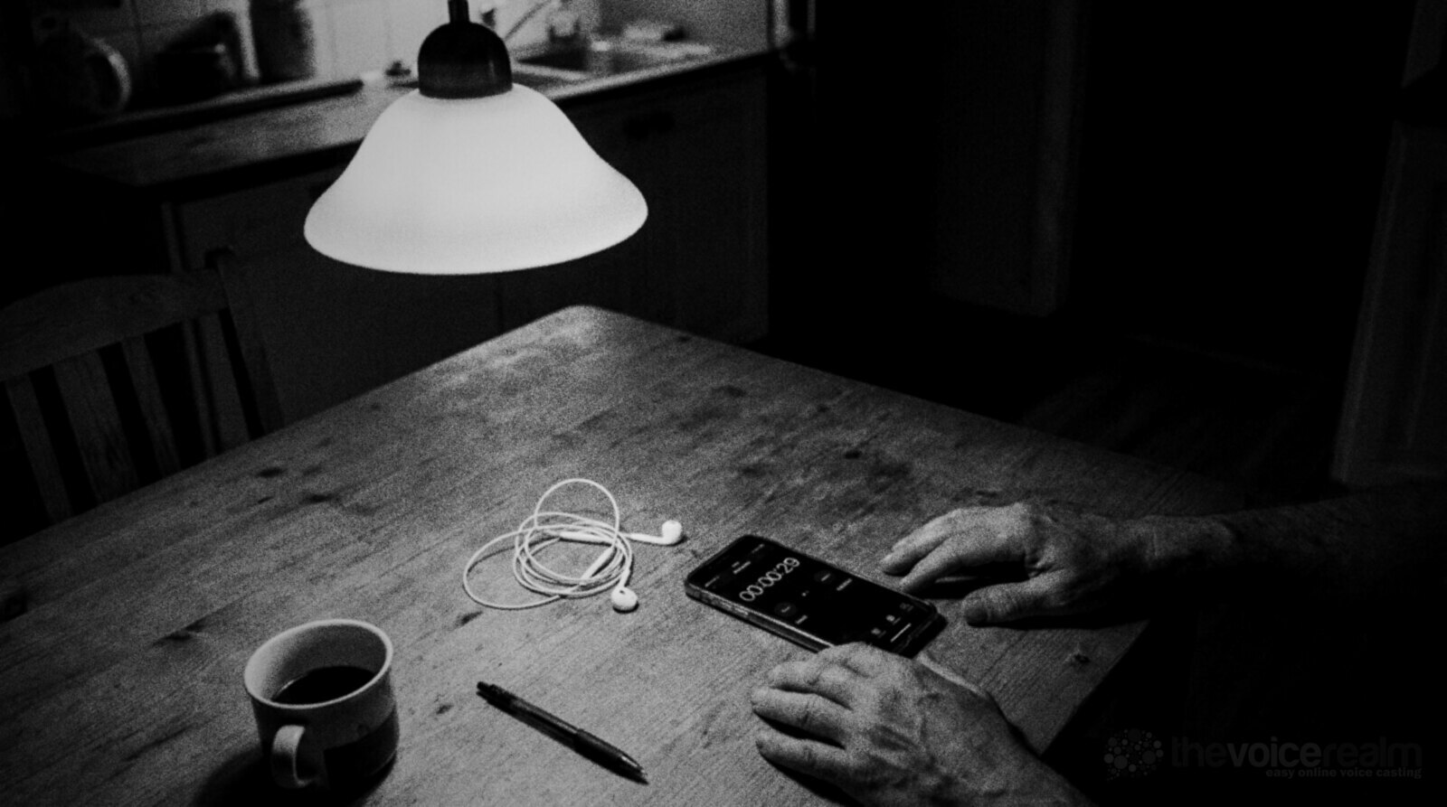 Black and white photo of a kitchen table late at night under a pendant lamp, a phone showing a stopwatch stopped at twenty-nine seconds, a tangle of wired earbuds, a pen, a mug of coffee and a man's hands resting on the table © The Voice Realm