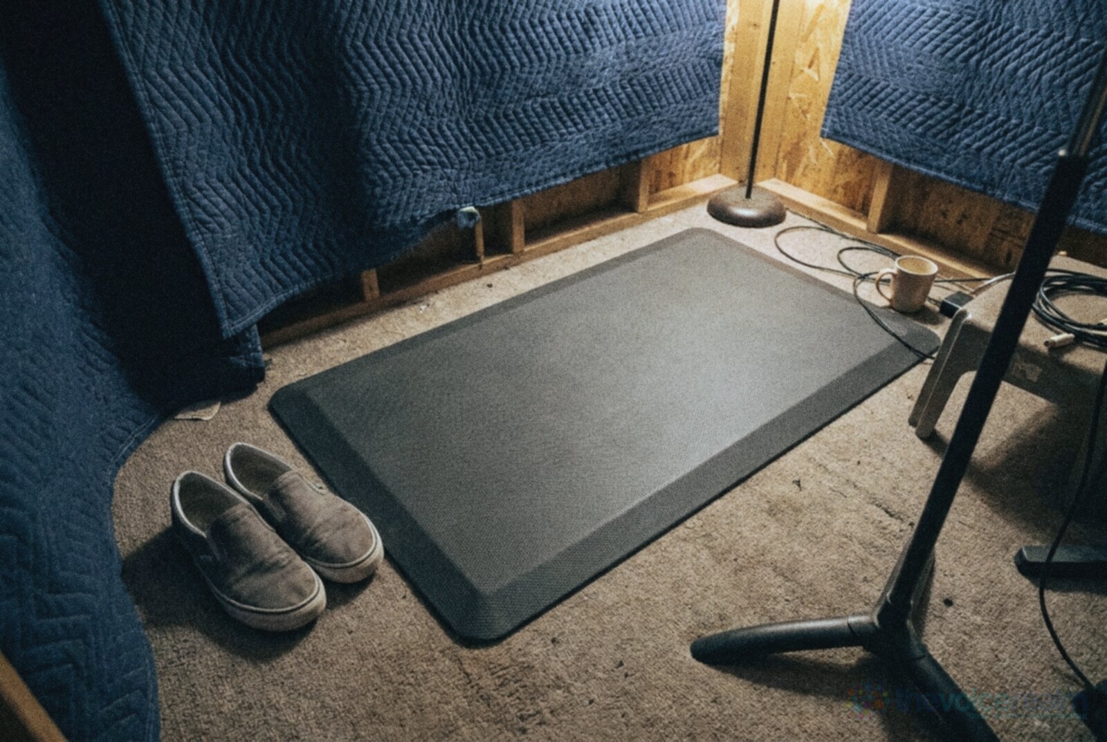 Looking down at the floor of a small home vocal booth lined with blue moving blankets: a grey cushioned standing mat on carpet, a pair of worn canvas slip-on shoes beside it, the legs of a stand and coiled cables, in cool grainy night film colours © The Voice Realm