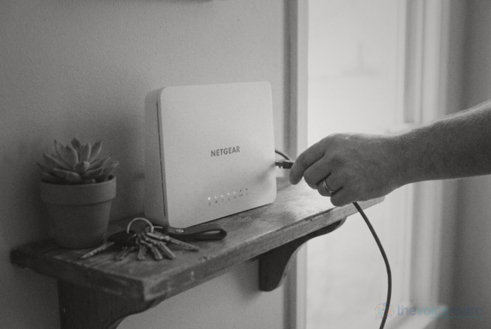 A hand plugging a network cable into a white home router on a wooden hallway shelf beside a set of keys and a small succulent, black and white photograph © The Voice Realm