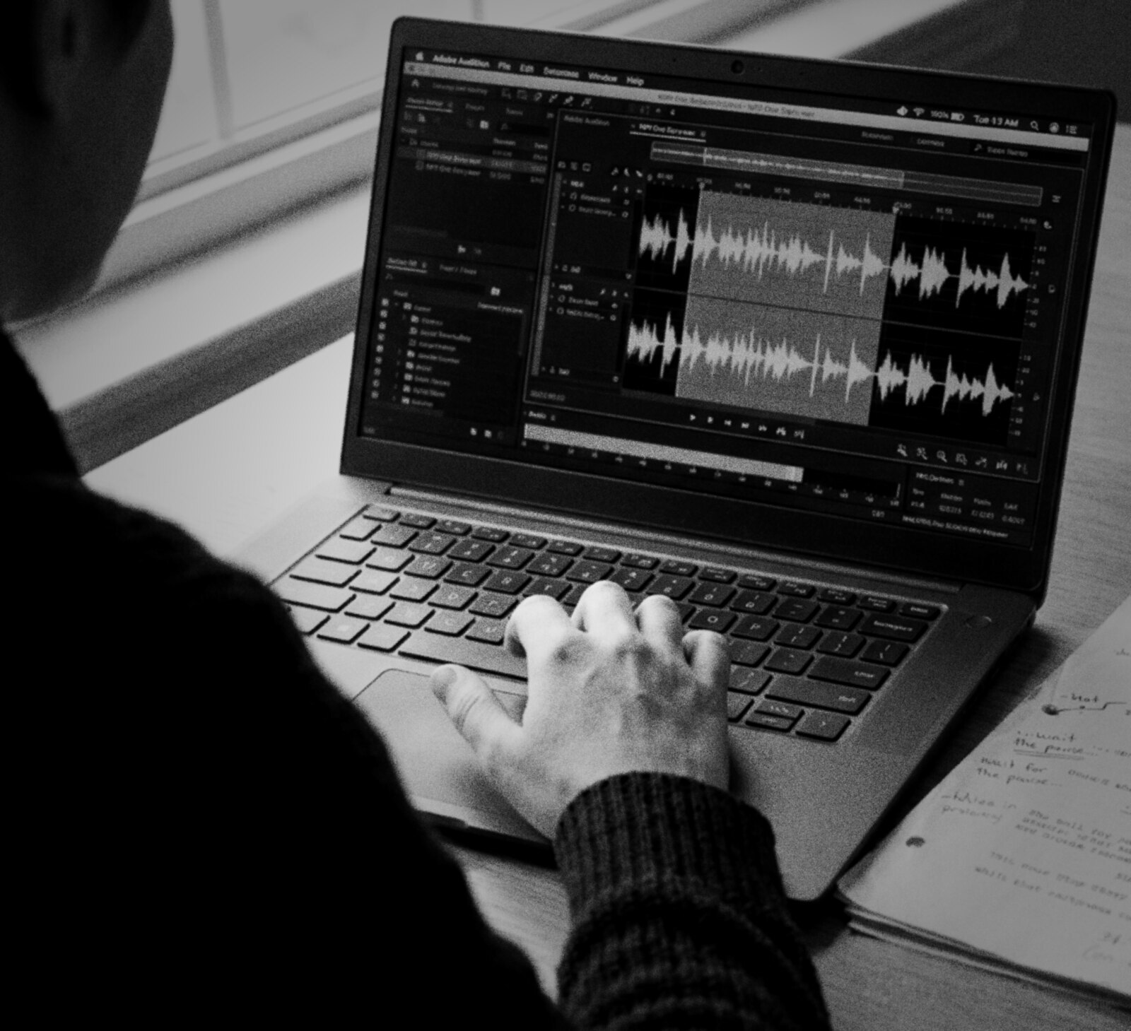 Over the shoulder of someone editing on a laptop, a highlighted section of a spoken word waveform in an audio editor on screen and their hand resting on the keyboard, black and white photograph © The Voice Realm