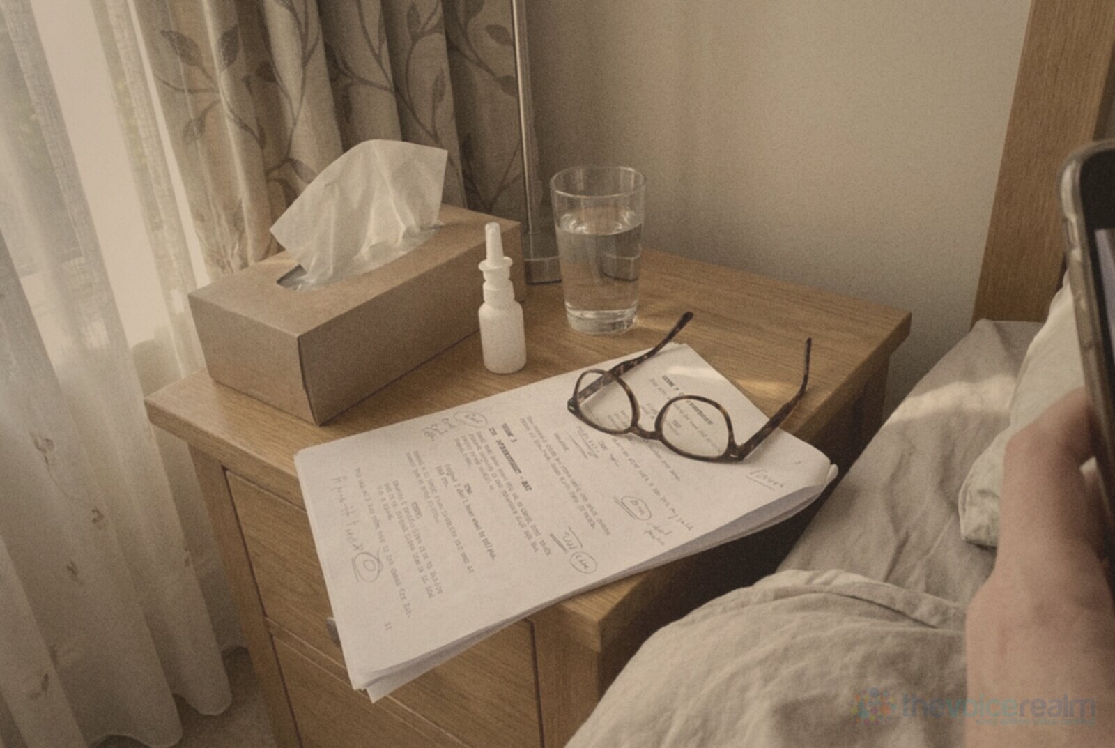 A bedside table in morning light with a box of tissues, a small plain white nasal spray bottle, a glass of water, tortoiseshell reading glasses and a marked-up script, a hand holding a phone at the edge of the bed, in faded 1970s print colours © The Voice Realm