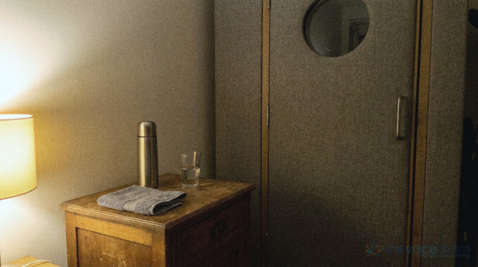 A thermos, a glass of water and a folded towel on a small wooden cabinet outside a padded recording booth door, lit by a lamp in the evening © The Voice Realm