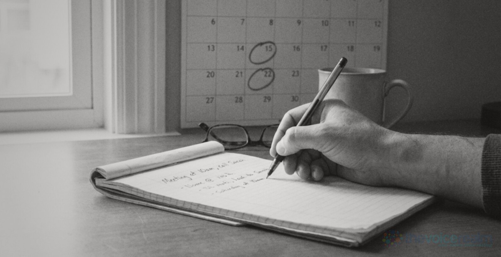 A hand writing with a pen in a paper notepad, a wall calendar with circled dates behind it, black and white photograph © The Voice Realm