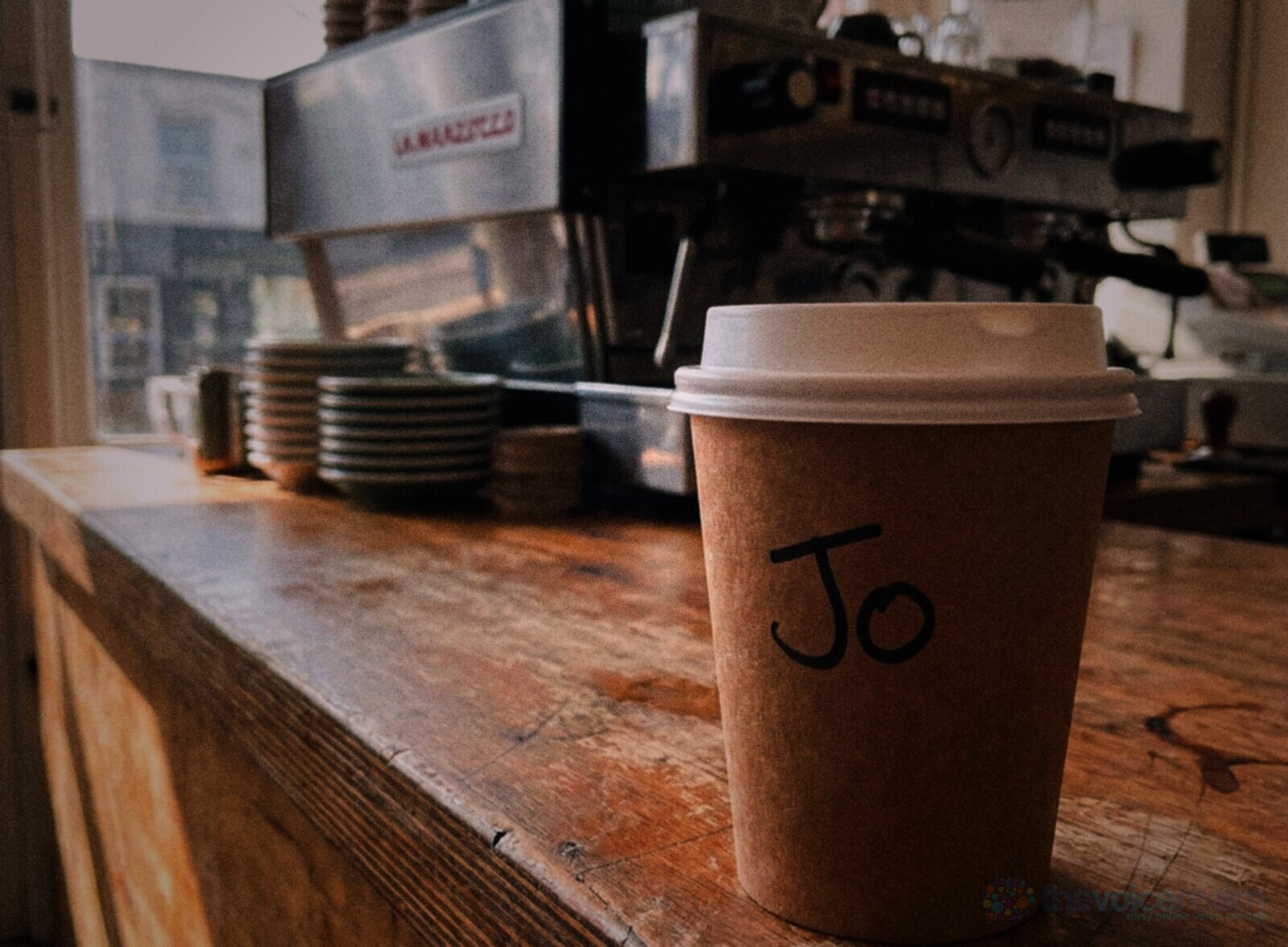 A takeaway coffee cup with the name Jo written on it in black marker, on a wooden cafe counter in front of an espresso machine © The Voice Realm