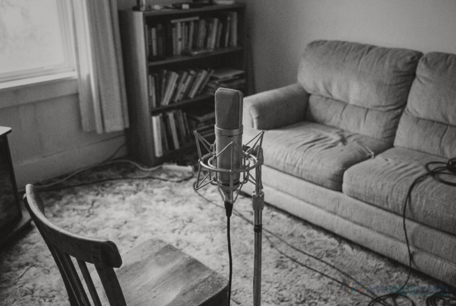 A large diaphragm condenser microphone in a shock mount on a stand in a carpeted living room with a bookcase and sofa, an empty wooden chair in the foreground, black and white photograph © The Voice Realm