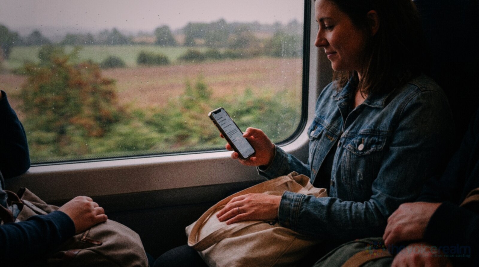 A woman on a train smiles as she reads an email on her phone, a canvas tote bag on her lap and rainy countryside passing the window, in rich film colours © The Voice Realm