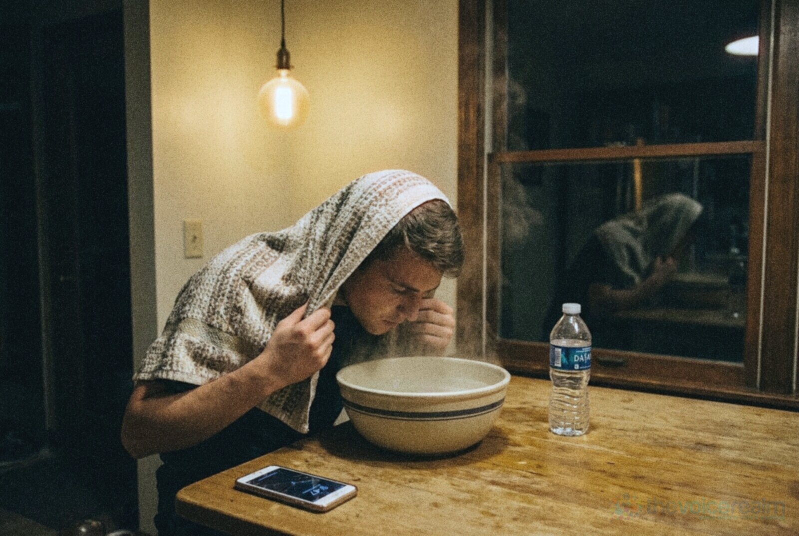 A young man with a towel over his head leans over a bowl of steaming water at a wooden kitchen table at night, a bottle of water and a phone beside the bowl under a warm hanging bulb, in cool grainy night film colours © The Voice Realm