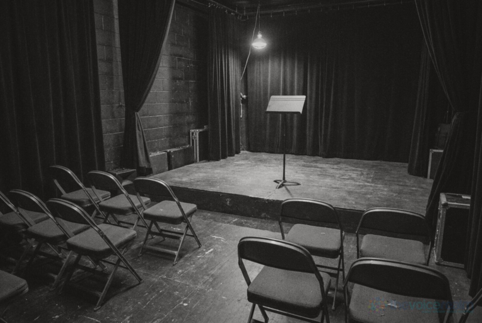 An empty black box theatre with rows of folding chairs facing a bare wooden stage where a single music stand stands under one hanging work light, black and white photograph © The Voice Realm